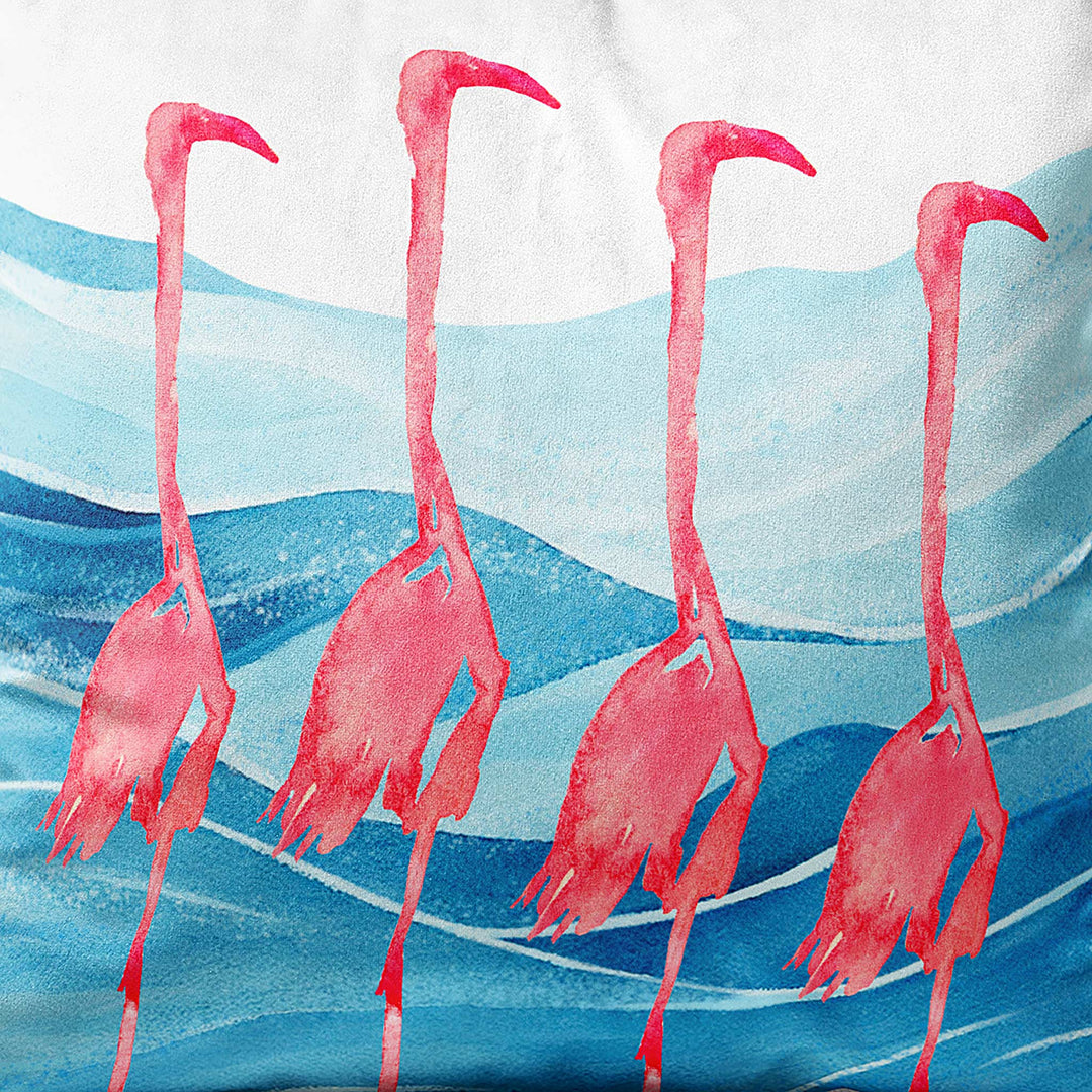 Close-up view of a square throw pillow featuring four watercolor flamingos standing in blue ocean waves, creating a coastal-inspired home decor accent.