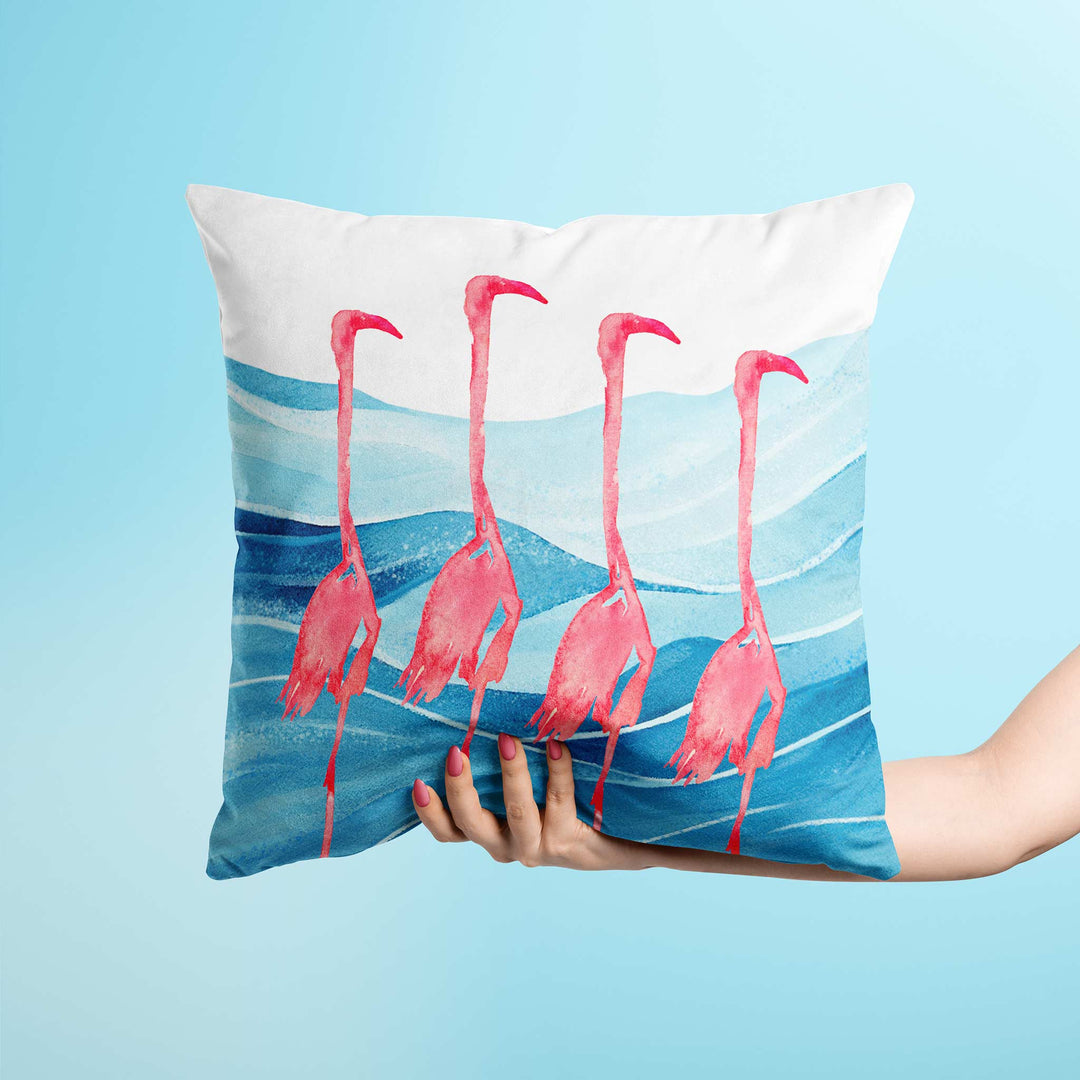 Close-up view of a square throw pillow featuring four watercolor flamingos standing in blue ocean waves, creating a coastal-inspired home decor accent.