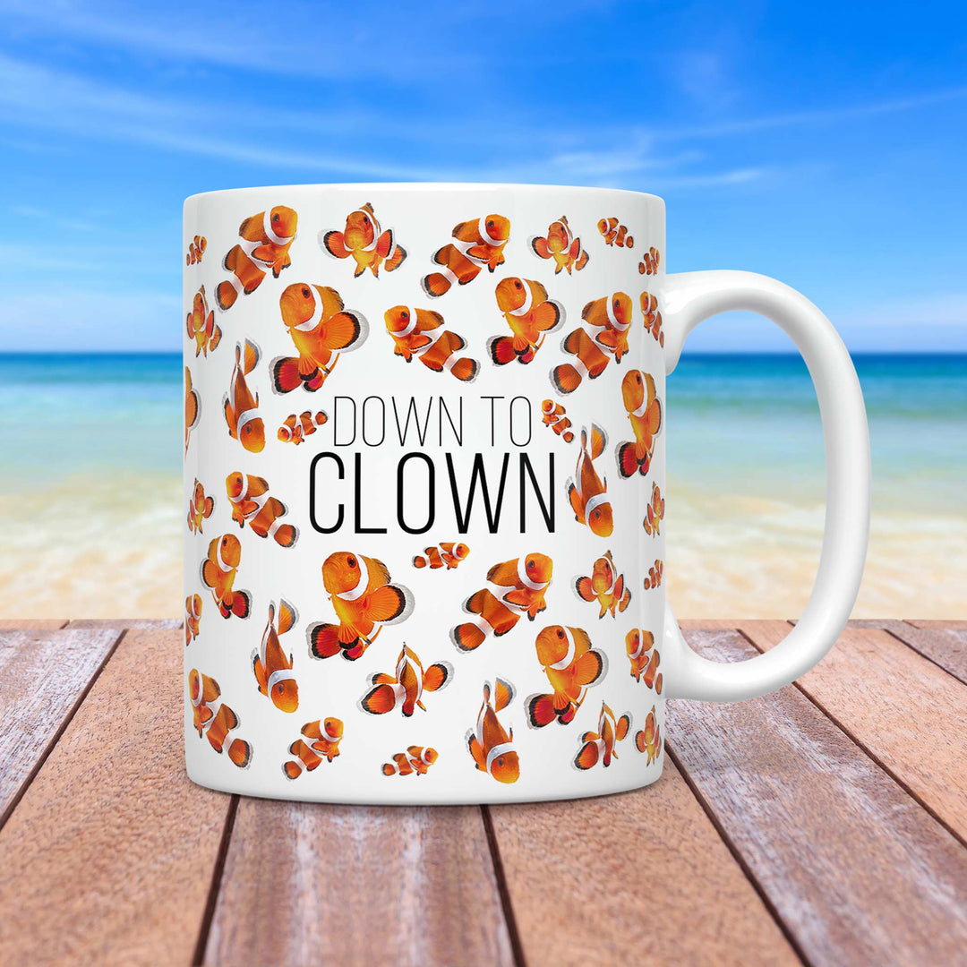 A white ceramic mug featuring a playful design with multiple clownfish illustrations scattered across the surface. The text “DOWN TO CLOWN” is centered in bold black font. The mug is placed on a wooden surface with a tropical beach scene in the background, showcasing a clear blue sky, ocean waves, and sandy shore.
