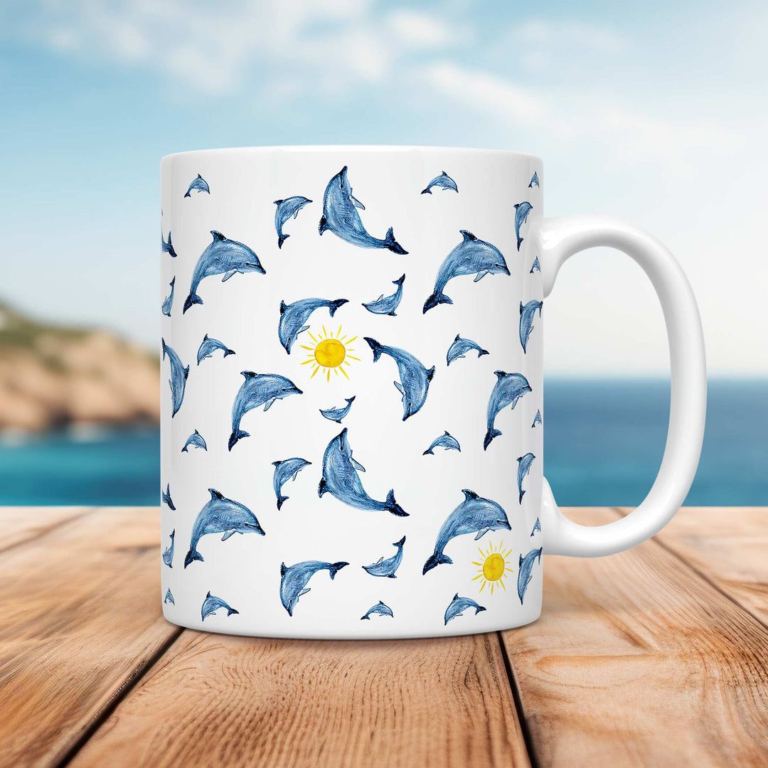 A white ceramic mug featuring a playful pattern of hand-drawn dolphins in various poses, leaping and swimming in different directions. Small yellow suns are scattered among the dolphins, adding a cheerful touch. The design has a watercolor-like texture with shades of blue. The mug is placed on a wooden surface with a coastal background, featuring a blurred shoreline, blue ocean, and soft sky.
