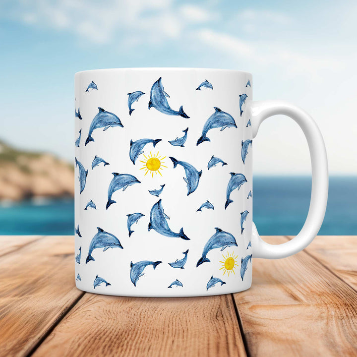 A white ceramic mug featuring a playful pattern of hand-drawn dolphins in various poses, leaping and swimming in different directions. Small yellow suns are scattered among the dolphins, adding a cheerful touch. The design has a watercolor-like texture with shades of blue. The mug is placed on a wooden surface with a coastal background, featuring a blurred shoreline, blue ocean, and soft sky.