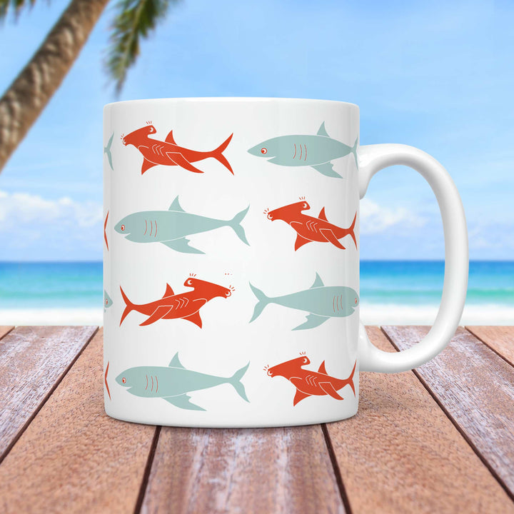 A white ceramic mug featuring a playful pattern of stylized sharks in two contrasting colors—light blue and burnt orange. The sharks have a minimalistic, cartoon-like design with simple line details and expressive eyes. The mug is placed on a wooden surface with a tropical beach background, including a palm tree, ocean waves, and a bright blue sky.