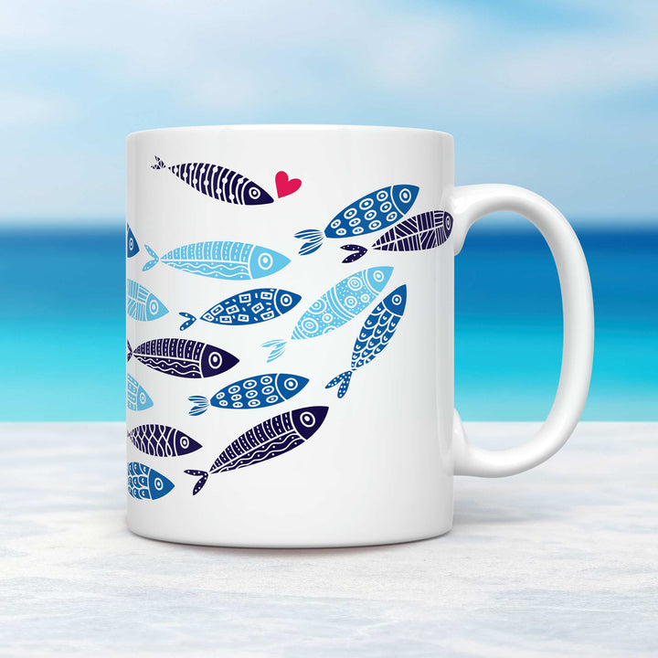 A white ceramic mug featuring a school of stylized fish in various shades of blue, each adorned with intricate geometric patterns. One fish stands out with a black and white striped design and a small red heart near its mouth. The mug is placed on a light-colored surface with a tropical ocean background, showcasing a gradient of turquoise waters and a soft blue sky.