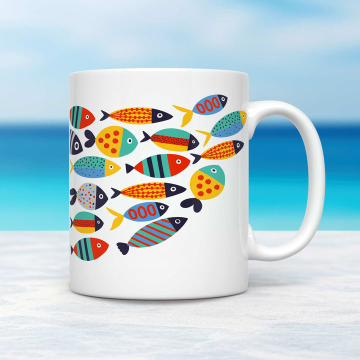 A white ceramic mug featuring a vibrant, modern design of stylized fish in bold, geometric patterns and bright colors, including red, blue, yellow, and green. The fish are arranged in a dynamic school formation, each with unique patterns like stripes, dots, and waves. The mug is placed on a light-colored surface with a serene tropical ocean background, showcasing clear blue waters and a soft sky gradient.