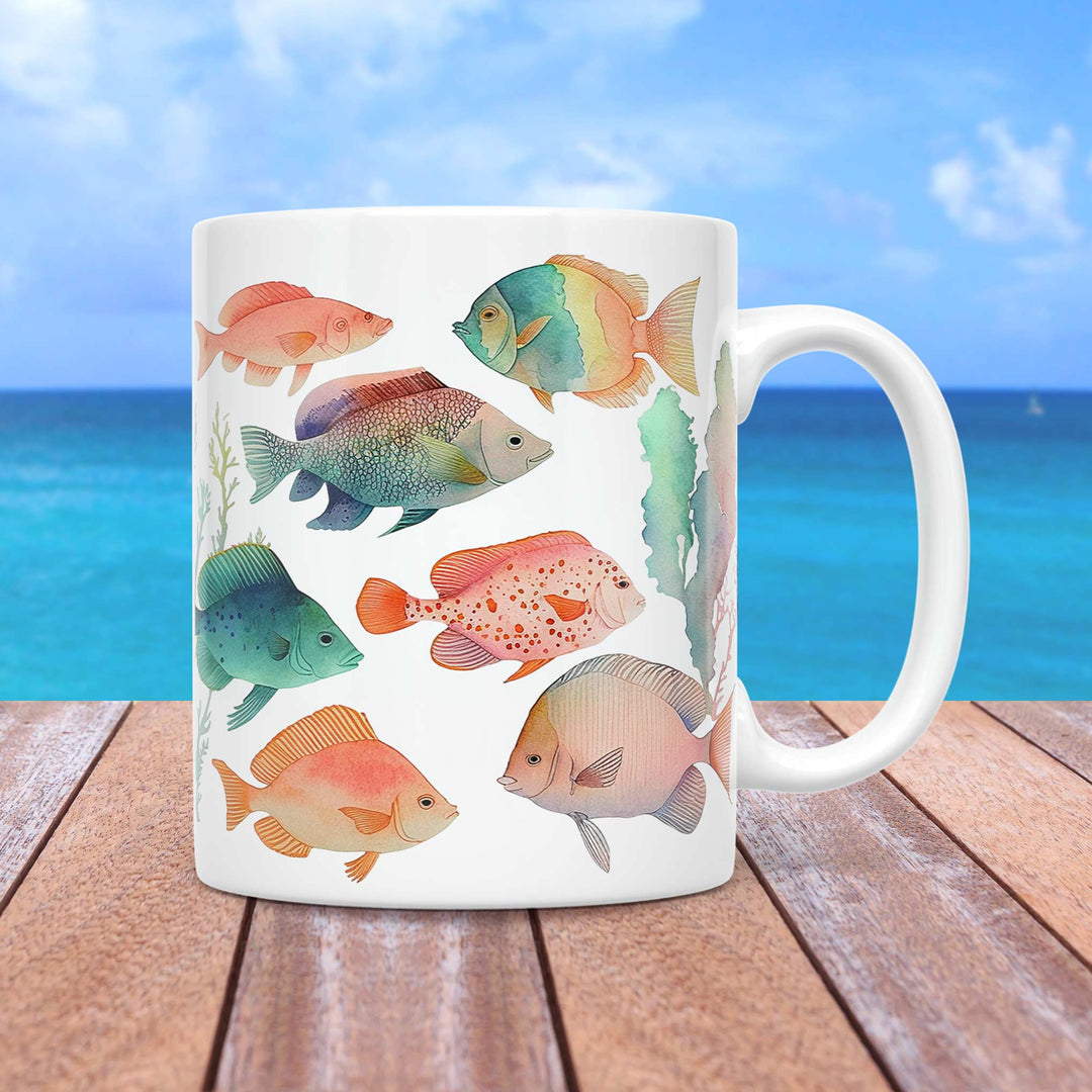 A white ceramic mug featuring a watercolor-style design of various tropical fish in soft pastel colors, swimming among delicate seaweed and coral elements. The fish display intricate details and a harmonious color palette of blues, greens, oranges, and pinks. The mug is set on a wooden surface with a tropical ocean backdrop, featuring a bright blue sky and turquoise waters.