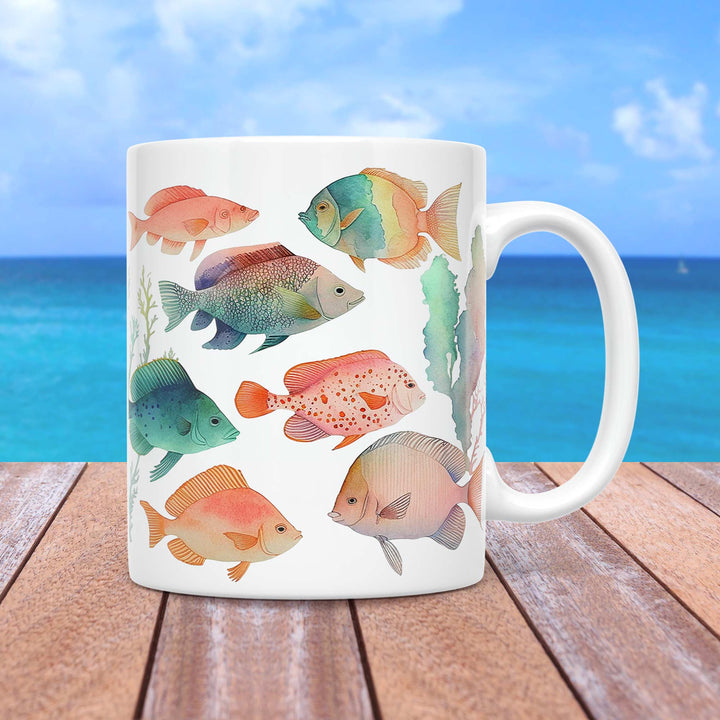 A white ceramic mug featuring a watercolor-style design of various tropical fish in soft pastel colors, swimming among delicate seaweed and coral elements. The fish display intricate details and a harmonious color palette of blues, greens, oranges, and pinks. The mug is set on a wooden surface with a tropical ocean backdrop, featuring a bright blue sky and turquoise waters.