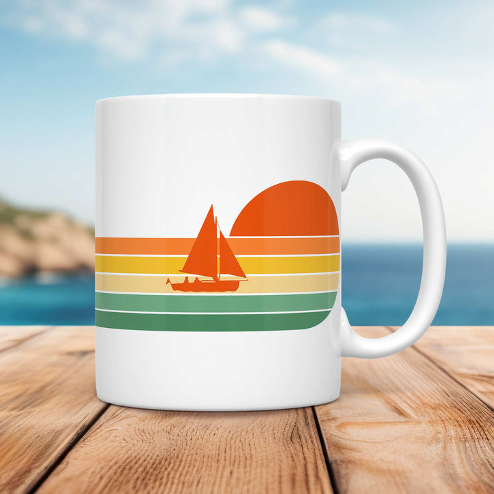 A white ceramic mug featuring a retro-style design of a sailboat silhouetted against a setting sun. The design incorporates warm, vintage-inspired horizontal stripes in shades of orange, yellow, and green, creating a nostalgic beachy vibe. The mug is placed on a wooden surface with a coastal background, showcasing a blurred shoreline, deep blue ocean, and soft sky.