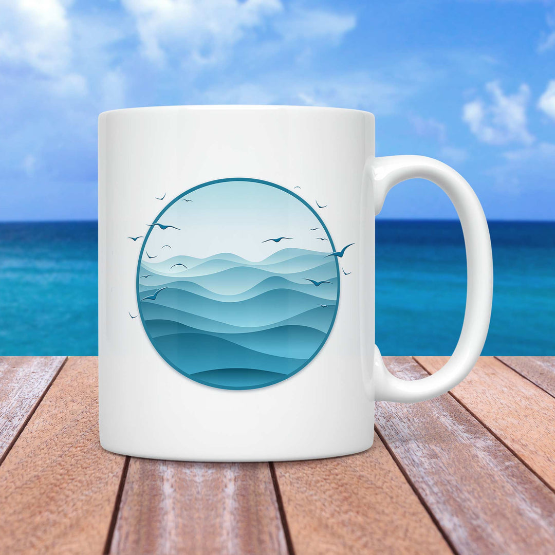 A white ceramic mug featuring a circular design of layered ocean waves in various shades of blue, creating a serene and minimalist seascape. Small silhouettes of birds are flying above the waves. The mug is placed on a wooden surface with a tropical beach background, featuring a clear blue sky and deep blue ocean.