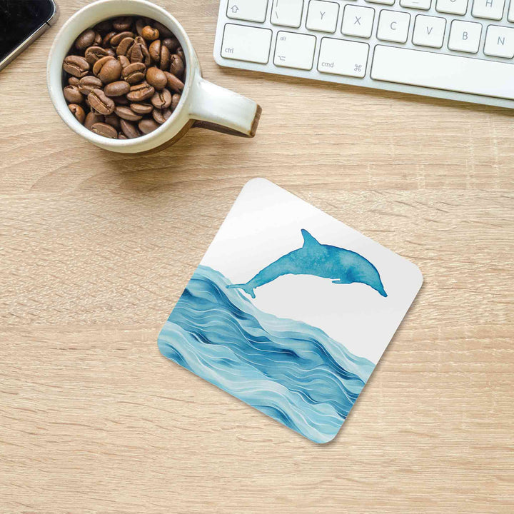 A dolphin-themed coaster featuring a watercolor-style dolphin leaping over ocean waves, placed on a light wooden desk. Nearby, a ceramic mug filled with coffee beans, a white keyboard, and part of a smartphone create a cozy and stylish workspace setting.