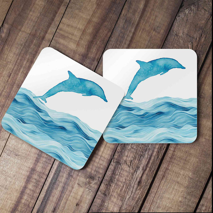 Two dolphin-themed coasters featuring a watercolor-style dolphin leaping over ocean waves, placed on a rustic wooden surface. The matching designs create a cohesive and stylish coastal-inspired decor piece.