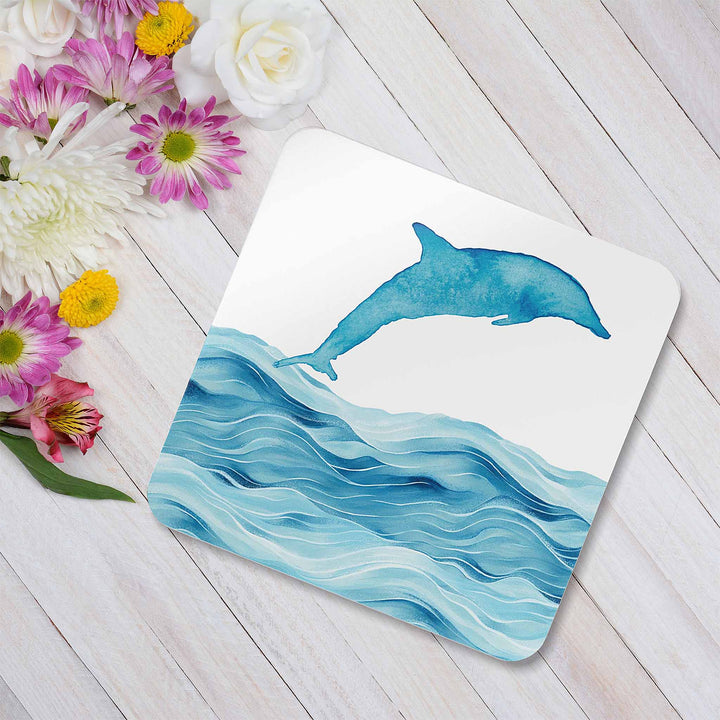 A dolphin-themed coaster featuring a watercolor-style dolphin leaping over ocean waves. The coaster is placed on a white wooden surface, surrounded by an arrangement of fresh flowers in shades of pink, white, and yellow, creating a soft and elegant coastal-inspired display.