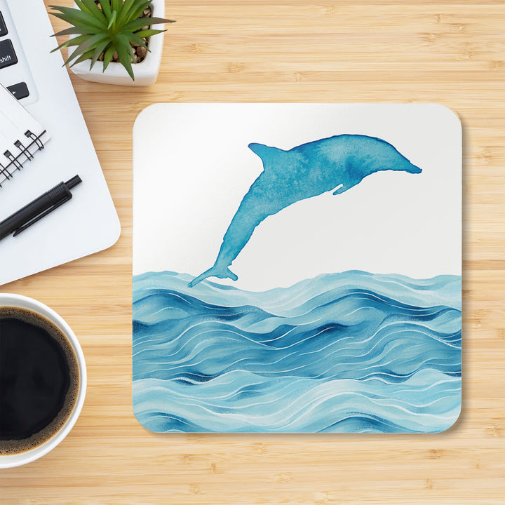 A dolphin-themed coaster featuring a watercolor-style design of a dolphin leaping over ocean waves. The coaster is placed on a light wooden desk alongside a cup of black coffee, a notepad, a pen, and a small potted succulent, creating a stylish and functional workspace setting.